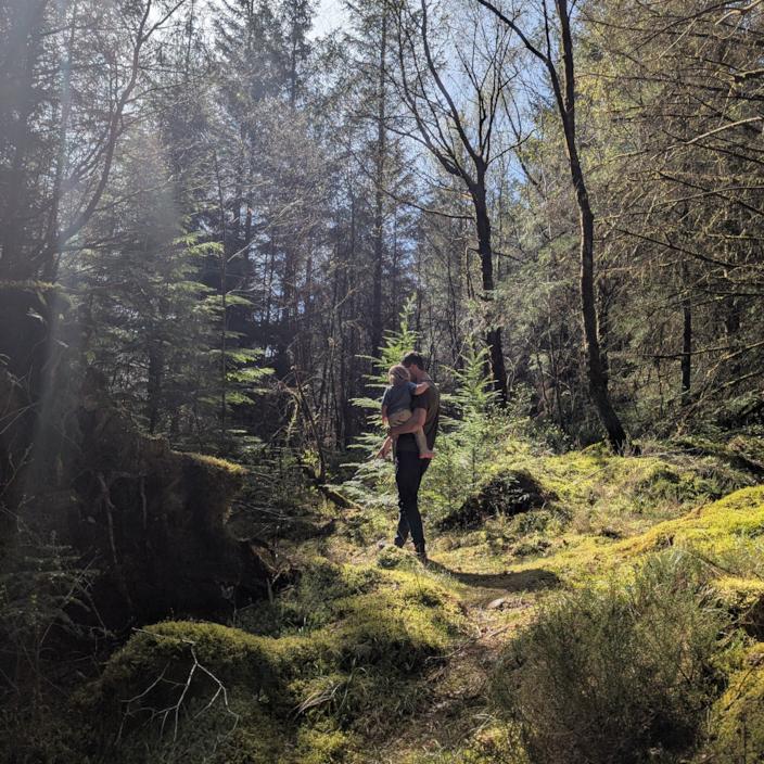 Foel in a mossy sunlit forest carrying a small child and pointing at something on the ground